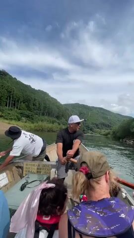The video shows a view from the back of a boat on a river, looking forward. Two men are in the boat, one at the front and one in the middle, both appearing to be guiding the boat with oars. The man in the middle is wearing a black t-shirt and grey shorts and is pointing with his right arm. He is also wearing a white baseball cap. The man at the front is wearing a white t-shirt with a black design and khaki shorts, and is also holding an oar. In the foreground, the backs of two people are visible. The person on the left has dark curly hair and is wearing a life jacket. The person on the right has blonde hair and is wearing a camouflage life jacket and a green baseball cap. The boat is moving down a green river, flanked by lush, tree-covered hills under a partly cloudy sky. There is text visible on the side of the boat in Japanese.