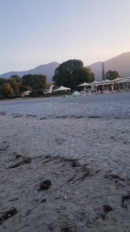 The video shows a beach scene at dusk or dawn. The sky is a clear blue with a hint of pink near the horizon. A single seagull is flying across the sky. The beach itself is made of pebbles and sand. In the background, there are trees and mountains. There are a few people on the beach, some walking near the water and others in the distance. There are also a few beach umbrellas and tents set up. The waves are gentle, lapping at the shore.