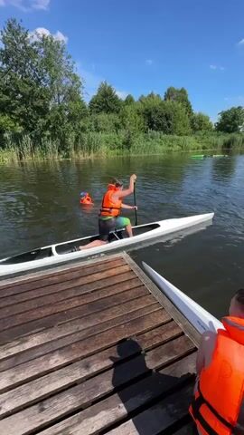 This video captures a sunny day on a calm body of water, likely a river or lake, with lush green trees and reeds lining the bank. The scene is set with a wooden dock in the foreground, two individuals in orange life vests are visible. One person is seated on the dock, their back to the camera, while another is in the water, wearing a blue swimming cap and an orange life vest, actively swimming.

In the water, a white kayak is being paddled by a person wearing an orange life vest and shorts. This individual is actively engaged in paddling the sleek kayak, suggesting a recreational activity or perhaps a lesson. In the background, further out on the water, another person is visible in the distance, also in the water and wearing an orange life vest. This suggests a group activity or a supervised session.

The weather appears to be fair and warm, indicated by the bright sunshine and the clear blue sky dotted with a few white clouds. The water is relatively still, reflecting the sky and the surrounding greenery, creating a peaceful and inviting atmosphere. The overall impression is one of outdoor recreation and enjoyment of a summer day. There is no visible text in the video.