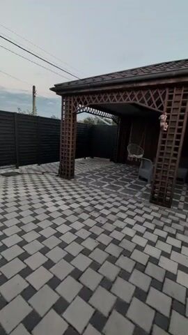 This media file shows a courtyard with a wooden gazebo and a paved area. The gazebo has a patterned roof and decorative lattice work on its sides. Inside the gazebo, there is a hanging chair and some outdoor furniture. The courtyard is paved with interlocking concrete blocks arranged in a diamond pattern. A dark fence runs along the left side of the courtyard, and a utility pole with electrical wires is visible in the background. The sky is overcast, suggesting a cloudy day. The overall impression is of a well-maintained outdoor living space. There is no visible text in the media file. The location context is Izmail, Ukraine.