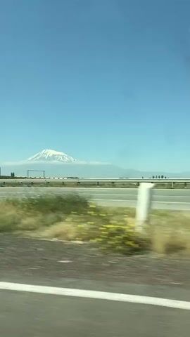 The video shows a large, snow-capped mountain in the background, partially obscured by clouds. In the middle ground, there is a highway with a metal guardrail. The foreground is blurred, suggesting the video was taken from a moving vehicle, possibly from a car window. The sky is clear and blue.