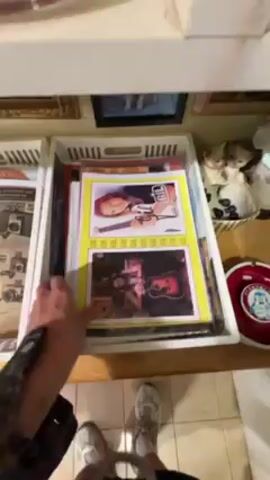 A person is shown removing items from a white plastic storage container. The container is sitting on a light brown surface, likely a table or shelf, and contains a variety of items including a magazine cover with the title "Splitting Heirs," a poster for Def Leppard's Greatest Hits Vault, and other papers. The shelf unit is also cluttered with items, including pictures in frames and decorative figurines. The person's arm and torso are visible, and they are wearing grey sneakers and dark pants.  The floor is tiled in beige and white.