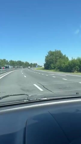 The video shows a view from inside a car on a sunny day, driving on a highway. The dashboard is visible in the foreground, and a dashboard ornament of a yellow rubber duck wearing a helmet and sunglasses with a propeller on top is positioned in the center of the frame. The duck is the primary focus, and the highway with green trees and blue sky is the background. The car appears to be in motion.