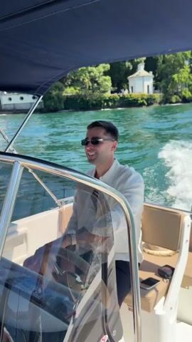 A man is steering a boat on a body of water. He is wearing sunglasses and a white shirt. He smiles and makes a peace sign with his hand. In the background, there are trees and a white building on the shore. The water is blue and there are white wakes behind the boat.