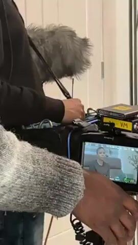 The video shows a woman with braided hair sitting on a gray couch. She is wearing a black long-sleeved shirt and gray jeans. Behind the camera is a sound man and a cameraman. The camera is a Blackmagic Design camera. The woman is being filmed.