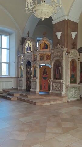 This video captures the interior of an Orthodox church in Ivashevo, Russia. The primary subject is a multi-tiered iconostasis, an ornate screen adorned with religious icons, which separates the nave from the sanctuary.

The iconostasis features numerous painted icons depicting saints and biblical figures, many with golden halos and backgrounds, set within intricately carved wooden or stucco frames. The central Royal Doors of the iconostasis are prominent, decorated with elaborate gold patterns and smaller iconic images. Other icons are visible on the side panels and upper tiers.

The church interior is bright, with light-colored walls and a vaulted ceiling. A large, decorative white chandelier hangs from the ceiling on the left side of the frame. The floor is tiled with a light, checkered pattern. Natural light streams into the space from a window visible on the left, indicating it is daytime.

The video starts with a static shot focusing on the right side of the iconostasis, then pans slightly to the right to reveal more of the church's depth, including another, larger iconostasis further back. It then zooms in on the details of the nearer iconostasis, showcasing its intricate artwork and craftsmanship. No people are visible, and no legible text can be discerned on the icons or other elements within the scene.