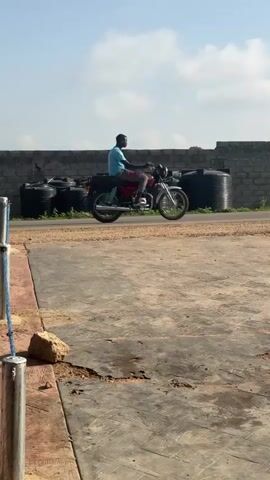 The video shows a man riding a motorcycle on a road, with a stack of black water tanks behind a gray brick wall in the background. The man is wearing orange pants, a purple shirt, and a cap. The motorcycle is black with silver accents. The road is made of gravel. The area in the foreground is made of cement. There is a rock on the ground next to a metal pole. The sky is blue with white clouds.