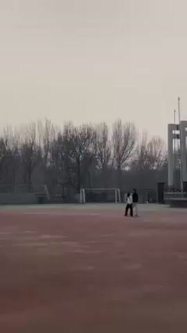 The video shows a sports field with a running track surrounding a grassy area, likely used for soccer.  In the first part, the sky appears overcast with a grey or hazy quality. Two people, a male and a female, walk together across the running track. The trees in the background are bare, suggesting it is winter or early spring. There is a soccer goal in the background. The second part of the video shows a group of people gathered around one of the soccer goals, with a soccer ball nearby.  Some of them appear to be warming up or preparing to play. The sky is brighter in this part, though the color is still muted.