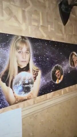 The video pans across a wall decorated with wallpaper that has a geometric pattern. On the wall, there is a large panoramic photograph or artwork depicting a woman holding a crystal ball against a backdrop of stars and a galaxy. Within the starry backdrop, there are circular cutouts or frames containing portraits of three other individuals. The camera then pans to the right, revealing a mounted deer head on the wall above the panoramic artwork. Further to the right, on the same wall, are three framed pictures. The first framed picture shows a pastoral scene with figures. The second is a framed poster or print with text and drawings. The third is a framed landscape painting of a field with trees. The video continues to pan, showing a staircase railing and a plant with white flowers in the foreground.