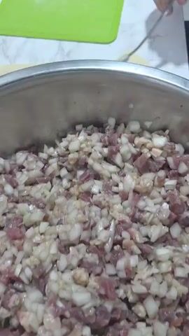 The video shows a close-up view of a person preparing dumplings. In the foreground, a stainless steel bowl is filled with a mixture of chopped meat and onions, the ingredients for the dumpling filling. A silver spoon is resting inside the bowl, with some of the mixture clinging to it. In the background, a person's hands are visible. The person is using the spoon to scoop the filling from the bowl and place it onto small, square pieces of yellow dough, which are arranged on a white surface next to a green cutting board. The action is focused on the process of filling the dumplings.