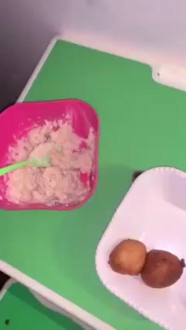 A top-down shot reveals a child's meal placed on a green table. In the upper left corner is a pink bowl with a mushy, light brown substance, presumably food, with a green spoon partially submerged in it. To the right of the bowl, a small, square, white decorative dish holds two small, spherical fried items that are different shades of brown. The table is mostly green with white trim.