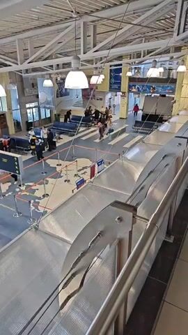 The video shows a high-angle view of what appears to be an airport waiting area. People are moving around, some with luggage. Some are sitting on benches. The floor has a large map of the world painted on it. There are several service counters, a shop, and some security barriers in the area. The space has a high ceiling with exposed metal beams and several pendant lights. The camera is positioned behind a transparent, curved barrier.