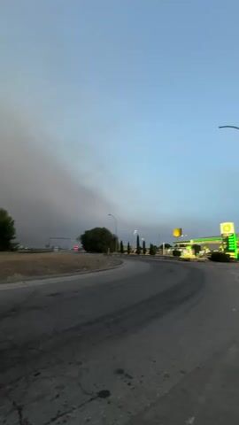The video shows a road with a roundabout and a gas station in the background. The sky is filled with smoke, indicating a fire nearby. The road is paved and appears to be well-traveled, with tire marks visible on the surface. There are streetlights and trees along the side of the road. The gas station has a yellow sign and several pumps. The overall impression is one of an industrial or suburban area affected by smoke.