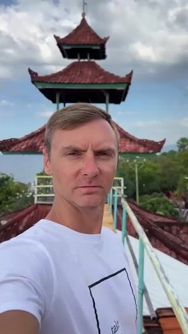 The video opens with a close-up of a man's face as he looks into the camera. He is wearing a white t-shirt with a black square design on it. Behind him, a red-roofed pagoda-style structure is visible, along with a wooden walkway. As the video progresses, the camera pans slightly to the left, revealing a scenic view of the ocean and lush greenery, including palm trees and flowering bushes. The man continues to look at the camera, occasionally winking or smiling. The background shows more of the tropical landscape, with glimpses of buildings and a swimming pool. The sky is partly cloudy, with white clouds against a blue sky. The overall impression is of a relaxed, outdoor setting with a man taking a selfie.