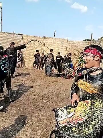 The video shows a cultural performance. Several people are dressed in traditional clothing and masks. Some people are holding whips. The background shows a partially constructed bamboo fence, a crowd of spectators, and a tent. The ground appears muddy or dusty. The sky is clear and blue.