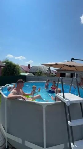 This is a vertical, eye-level, medium shot of a family enjoying a sunny day in a backyard above-ground pool. The pool has a grey exterior and a blue interior filled with clear water. A white ladder with grey rungs leads into the pool on the right side. A large, tilted, beige umbrella with a black pole provides shade over a brick structure and a portion of the pool. In the background, a green fence runs along the perimeter of the yard, with houses and trees visible beyond it under a bright blue sky with a few scattered white clouds.

In the pool, there are five people. On the left, a man with tattoos on his arm is partially submerged, leaning back on a bright green inflatable. He is looking to his right. To his right and further into the pool, a young boy in a blue swim shirt is playing with a yellow and blue water gun. He is looking at the man. Further back and to the right of the man, two young girls are in the water. One, in the center, is wearing a pink top and is holding a blue inflatable ring. The other, to her right, is wearing a pink top and appears to be in a blue inflatable. To the far right of the pool, a woman with blonde hair is sitting on the edge of the pool, partially submerged, looking towards the left. Another young child is on the right side of the pool, near the ladder. The atmosphere is relaxed and playful, with the water guns suggesting a game is in progress.