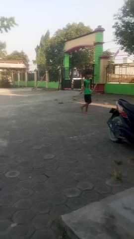 The video shows an outdoor scene in what appears to be a schoolyard or a public area. The ground is paved with hexagonal grey stones. In the background, there is a large, colorful archway serving as an entrance to a property, painted yellow, red, and green. Behind the archway, there are black metal gates and a fence. To the left of the entrance, there is a building with a light-colored wall and a green fence. To the right of the entrance, there is another fence and some trees. A young person wearing a green t-shirt and dark shorts is walking towards the right side of the frame. A dark-colored motorcycle is parked on the right side of the paved area, close to the viewer. The sky is bright and clear, suggesting it is daytime. The overall impression is one of a typical sunny day in an outdoor public space.