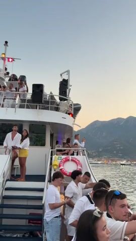 A boat is moving through the water near a coastline with mountains in the background. Several people are visible on the boat, most of whom are wearing white clothing. Some are standing, looking out at the scenery or interacting with each other. Others are seated or leaning on the railing. There are speakers and other equipment on the upper deck of the boat. The sun appears to be setting, casting a warm glow on the sky and water. Other boats and buildings are visible in the distance along the coast.