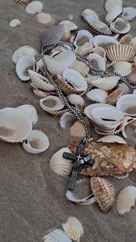 A close-up shot focuses on a collection of seashells scattered across a sandy surface. A silver chain with a cross pendant is draped over some of the shells. The cross is dark, with an ornate design and text inscribed on its surface, but the text is too small to be legible. The shells vary in size, shape, and color, ranging from small, white, ribbed shells to larger, cream-colored ones with darker markings. Some shells are broken, while others appear intact. A piece of weathered, brown, and tan material, possibly a larger shell or rock, is also visible amidst the smaller shells. The lighting suggests it may be daytime, with the sand appearing damp in places. The overall scene evokes a peaceful, natural setting, reminiscent of a beach. There are no people visible in the frame. The environment, with its sandy ground and seashells, strongly suggests a coastal or seaside location. Given the location context of Sihanoukville, Cambodia, the scene is likely a beach. The arrangement of the shells and the presence of the pendant could suggest a moment of reflection or a found treasure.