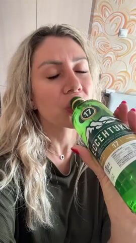 A blonde woman with her eyes closed is drinking from a green bottle of Essentuki No. 17 mineral water. She is wearing a dark green ribbed top and a delicate necklace. The background shows a beige and orange patterned wallpaper and white cabinetry. Her right hand holds the bottle to her lips, and her fingernails are painted red.
