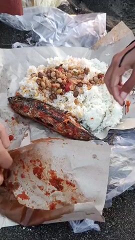 The video begins with a close-up of a meal laid out on paper on a dark, sandy beach. The meal consists of white rice, a whole grilled fish, and a topping of what appears to be beans and other ingredients. There are also plastic bags and crumpled paper scattered around the food. A person's hand is seen reaching for the rice. The camera then pans out slightly to reveal two individuals sitting on the beach. The person on the left is wearing a maroon outfit and is partially visible from the waist down, with their feet exposed. The person on the right has their arm and hand visible. As the video progresses, the camera shifts to capture a wider view of the beach and the ocean. The sun is setting, casting a warm glow over the scene, with a few distant boats visible on the water. The overall atmosphere suggests a casual meal enjoyed by the sea.