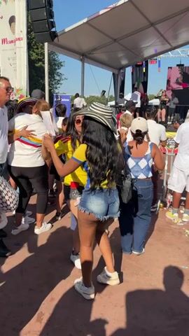 Two women in matching yellow and blue tops and denim shorts are dancing together outdoors. One woman wears a wide-brimmed black and white striped hat. They are holding hands and appear to be moving in sync. A stage with a large screen and sound equipment is visible in the background, along with other people watching the performance. The ground is covered in a reddish-brown dirt or gravel. The lighting suggests it is daytime, with shadows indicating the sun is present. The overall atmosphere appears to be festive, possibly at an outdoor festival or concert.