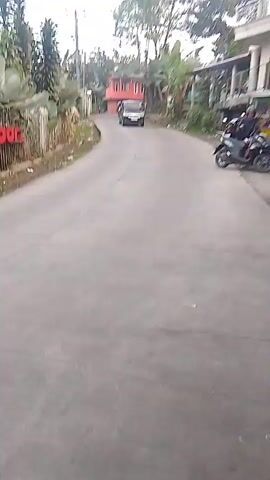 A video shows a paved road in a rural or suburban area. A black van is driving towards the camera. To the right of the road, a person on a motorcycle is parked. There are buildings and trees on either side of the road. The sky is overcast. The video appears to be filmed from a moving vehicle.