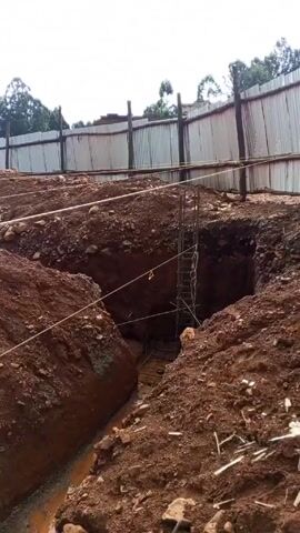 The video shows a building site with excavated trenches in red soil. There are metal rebar grids placed in some of the trenches, presumably for concrete reinforcement. White string lines are stretched across the site to guide construction. A red shovel lies in one of the trenches. In the background, a corrugated metal fence surrounds the site, with trees visible behind it. The sky is overcast. There is no visible human activity in the video.