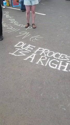 The video shows ground-level footage of people standing on asphalt on which chalk drawings and writings have been made. Messages written in white chalk include "DUE PROCESS IS A RIGHT" and "HUMAN OVER $$$". The chalk writings extend beyond these messages, with additional writings and doodles rendered in green chalk. Several individuals are visible from the waist down, their feet and lower legs present in the frame. They are wearing a variety of shoes, including brown loafers with an animal print, brown boots, black shoes, and blue sneakers. In the background, a sign is partially visible, but the text is illegible. The scene suggests a demonstration or protest, with chalk messages reflecting values and principles.