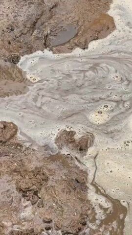 A close-up aerial view shows muddy ground with swirling white foam. The foam moves in circular patterns, creating a marbled effect on the surface of the water. The mud is a dark brown color, and there are some small rocks and debris visible within it. The overall impression is one of a natural, earthy scene.