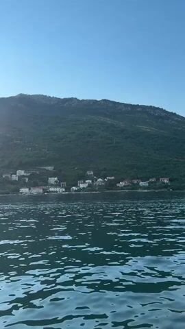 The video captures a serene scene from a boat on the water, looking towards a coastal village nestled at the foot of a lush, green mountain. The water in the foreground is slightly choppy, with sunlight glinting off its surface, creating a dynamic play of light and shadow. In the distance, a line of white and red-roofed houses forms a picturesque settlement along the shoreline. The mountain behind the village is densely forested, with a few scattered structures visible on its slopes, suggesting a rural and tranquil environment. The sky is a clear, bright blue, indicating fair weather. The angle of the sun, casting a soft glow and a subtle lens flare on the left side of the frame, suggests it is either early morning or late afternoon. The overall mood is peaceful and picturesque, evoking a sense of calm and natural beauty. There is no visible text in the media file. The location is Đurići, Montenegro.