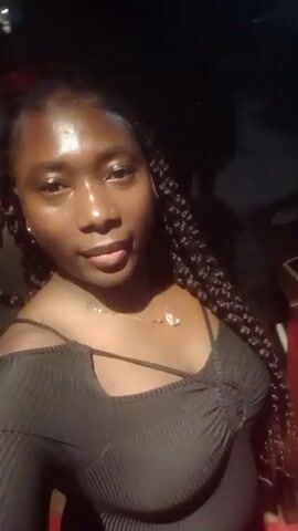 The video shows a woman in a black off-shoulder top with spaghetti straps and long braids. She is wearing a gold necklace with a small pendant and a nose ring on her left nostril. She is inside, possibly at night, with limited lighting. The background is dark, with glimpses of a red surface and some objects that appear to be a table and a phone. The woman looks directly at the camera and speaks briefly, her expression shifting from a neutral gaze to a small smile. The camera appears to be handheld and stable.