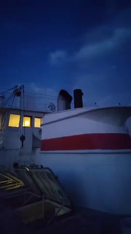 The video shows the side of a white boat with a red stripe at dusk. The sky is a deep blue with a few wispy clouds. There are illuminated windows on the boat, and what appears to be two black smokestacks on the boat. Some additional equipment is visible on the top of the boat, but it is difficult to discern the details due to the darkness. The camera is still and captures the scene from a low angle. A loud noise is heard at the end of the clip.