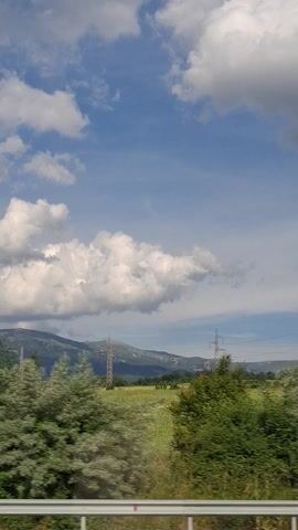 This video captures a view from a moving vehicle, likely a train, traveling through a rural landscape in Studena, Bulgaria. The perspective is tilted, giving the impression of a sideways view. Dominating the scene is a vast expanse of blue sky filled with dramatic, billowing white clouds.

Stretching across the frame are several high-voltage power lines, supported by tall, lattice-style metal pylons. These pylons, visible against the sky, indicate the presence of significant electrical infrastructure in the area.

To the right of the frame, a vibrant green landscape unfolds. Lush trees and bushes line the edge of the frame, transitioning into a grassy field that appears to be a meadow or farmland. In the distance, a range of rolling hills or mountains is visible, partially shrouded in a light haze, suggesting a considerable distance.

The time of day appears to be daytime, with bright sunlight illuminating the clouds and the landscape. The weather seems pleasant, with a mix of sun and clouds, typical of a summer day. There is no visible text in the video. The overall mood is one of travel and observing the natural and man-made elements of the Bulgarian countryside. The scene is peaceful and showcases the open, scenic terrain.