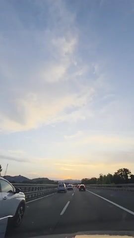 The video is a vertical shot from inside a vehicle on a highway at dusk. The sky is a pale blue with scattered clouds, transitioning to a soft yellow and orange near the horizon where the sun is setting behind mountains. Several cars are driving on the highway in the same direction as the camera. In the foreground, the side of a white SUV is visible on the left. The road has white lane markings.