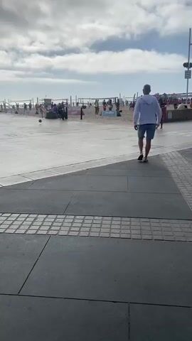 The video shows a beach scene with a paved walkway in the foreground. A man in a white hoodie and blue shorts walks away from the camera on the walkway. Further in the background, people are playing beach volleyball on the sand. There are volleyball nets set up, and spectators are visible. The sky is cloudy with some blue patches. To the right of the walkway, there is a traffic signal with a red light illuminated. A woman with a surfboard walks towards the camera on the walkway.