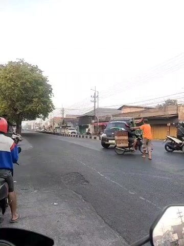 A black pickup truck with a green bed is turning right across traffic. A man in an orange shirt is walking across the road, presumably assisting the truck. A motorcycle is stopped in the road next to the truck, and another motorcycle is behind it. Two people are riding on a white scooter, and another person is riding a motorcycle behind them. A person on a motorcycle with a red helmet is on the left side of the road. Buildings line the right side of the road, and a large tree is on the left. The sky is overcast.