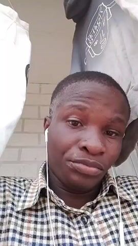 A person with dark skin and short hair, wearing a plaid shirt and white wired earbuds, is looking into the camera. The person’s expression changes from slightly smiling to a serious expression, then to a small smirk. Behind them, there is a white brick wall and clothes hanging.