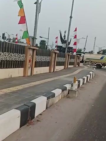 A video shows a street scene with a sidewalk and a road. A large monument with flags is visible in the background. A white tanker truck is driving on the road. The weather appears to be overcast. The video is shot from a moving perspective, suggesting it was recorded from a vehicle.