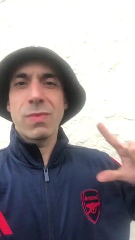 A man with dark hair and a light complexion, wearing a black bucket hat and a navy blue Adidas tracksuit jacket with an Arsenal logo on the left chest, faces the camera. He initially makes a scrunched-up facial expression, then relaxes into a smile, makes a peace sign with his right hand, and subsequently gestures with his right index finger as he chants "Arsenal" multiple times. A plain, light-colored textured wall is visible in the background.