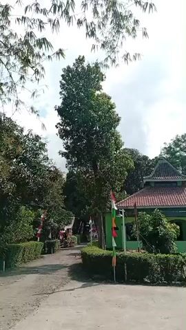 The video shows a path leading past a green building with a red tiled roof and a white dome. The building appears to be a place of worship. There are trees and bushes around the building. Several flags are visible along the path, including what appears to be the Indonesian flag. The sky is cloudy. The video is taken from a moving perspective, suggesting it might be from a vehicle or someone walking. The overall scene is one of a rural or suburban environment, likely in Indonesia, given the flags.