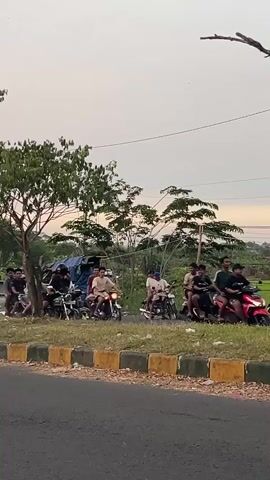 A group of people are gathered on motorcycles on the side of a road. The sky is overcast and the sun appears to be setting. There are trees and some structures in the background. The people are of various ages and are casually dressed. Some of the motorcycles are parked, while others appear to be in motion. The road is paved and has a yellow and black curb. The overall atmosphere appears to be relaxed and social.