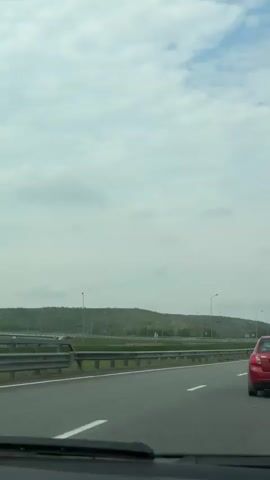 The view from inside a car driving on a multi-lane highway. Another red car is in the right lane ahead of the viewer's vehicle. A green, grassy hill is visible in the distance, and streetlights line the side of the road. The sky is overcast with a mix of white and gray clouds. The car is moving smoothly, with the highway lines appearing clear and well-maintained.