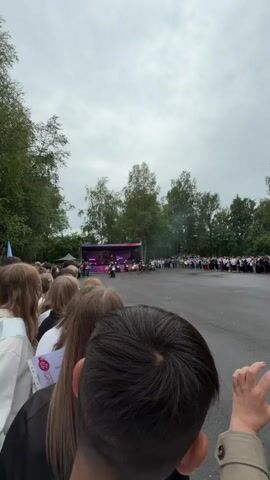 The video shows a crowd gathered in an outdoor area with a stage and trees in the background. Several motorcycles are performing stunts in front of the stage, with smoke visible. People are standing behind a barrier, watching the performance. One person in the foreground is holding a document with Russian text. The sky is overcast.