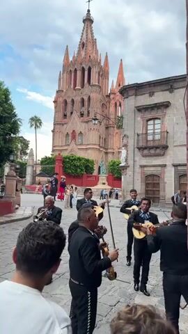 A mariachi band performs in front of a towering church in San Miguel de Allende, Mexico.  The band is comprised of about eight men dressed in black mariachi suits with silver embroidery. They play a trumpet, violins, and various guitars. A few people are gathered around watching and filming the performance.  In the background, the Parroquia de San Miguel Arcángel is visible. The weather is partly cloudy. The ground is cobblestone.  People walk in the background, some dressed in traditional attire, likely attending an event.