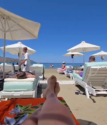 The video shows a first-person perspective of someone lying on a beach towel on a sandy beach, looking towards the ocean. There are several lounge chairs with umbrellas on the beach. In the foreground, the person's legs and feet are visible. In the mid-ground, people are seen lying on lounge chairs, walking on the beach, and swimming in the ocean. The ocean is blue and calm, and the sky is clear and blue. There are some hills or mountains in the background. The scene appears to be a sunny day at a beach resort.