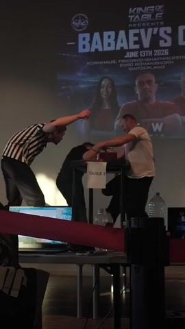 An arm wrestling match is underway. A referee in a black and white striped shirt stands on the left, arms raised. Two competitors are seated at a table labeled "TABLE 2". The competitor on the left, wearing a black shirt, is leaning forward, arm on the table. The competitor on the right, wearing a white t-shirt, is also leaning forward, arm on the table, and appears to be winning. A large screen behind the table displays text that includes "KING OF TABLE", "BABAEY'S CUP", and a date "JUNE 13TH 2026", along with a location in Switzerland. The environment appears to be indoors, possibly a venue or hall, with lighting focused on the arm wrestling table.