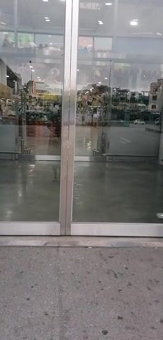 The video captures the opening of automatic glass doors at the entrance of a large supermarket or retail store in Ha Tu Ward, Vietnam.

At the beginning, the two main glass doors, framed in silver metal, are closed. The polished concrete floor in front of the doors reflects the bright overhead lighting from within the store. Through the clear glass, the interior is visible, showing a spacious retail area with numerous display shelves and products. Colorful decorative items, specifically umbrellas and beach balls, are suspended from the ceiling, suggesting a festive or seasonal theme, possibly summer. Reflections on the glass also reveal an outdoor area, possibly a parking lot or street, with distant buildings under an overcast sky or during the daytime.

As the video progresses, the automatic doors slide open smoothly, revealing the full extent of the store's entrance. Two metal security gates (anti-theft systems) flank the now-open doorway. Inside, a fresh produce section is immediately visible on the left, stocked with items like watermelons displayed in red crates. Further into the store, long aisles filled with various packaged goods and merchandise can be seen, all brightly lit by fluorescent lights. The store's interior floor is also a reflective, light-colored surface. No people are clearly visible in the immediate vicinity of the entrance as the doors open, but the setup implies customer entry. The store appears clean, well-organized, and spacious.