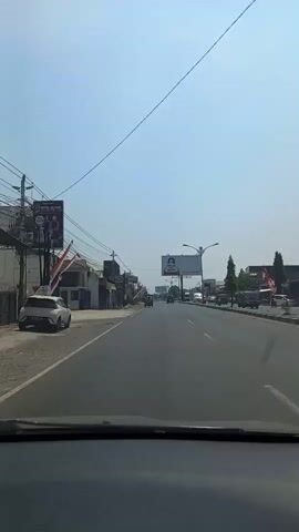 A first-person view from a vehicle shows a paved road with vehicles and buildings on either side. The sky is clear and blue. A large billboard with text and images is visible in the distance. Traffic lights and utility poles are also present. The video appears to be recorded during the daytime.