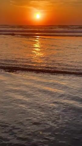 The video shows a sunset over a beach. The sun is a bright orange circle low on the horizon, casting a golden reflection on the water. Gentle waves are rolling in towards the shore, creating ripples and foam on the surface. The sky transitions from a warm orange near the sun to a softer yellow and pink further away. The overall scene is peaceful and serene. There are no people or objects visible in the video, just the natural landscape.